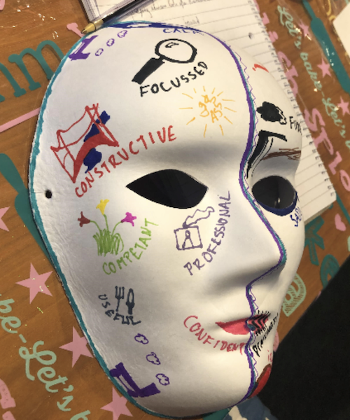 A white paper mask that has been decorated with text and drawings sitting on some brown paper with a notepad and pen in the background. The right side of the mask has positive words like focussed – with a picture of a magnifying glass; constrictive - with a drawing of a bridge; competent - with a drawing of some flower; useful with a drawing of a for and knife; professional - with a drawing of an ID badge; confident is written under the mouth which is painted red. On the left side there is a drawing of a black cloud with some words which are obscured.