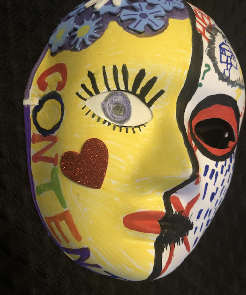 A paper mask which has been decorated by hand. The right side of it is yellow; it has blue and purple paper flowers as hair; the eye is blue, with long black eyelashes; it has a red love heart on the cheek; its lips are read; the word content is written down the side in different coloured letters. The left side is white; there is a house drawn on the forehead in blue and also a question mark; the eye is dark and circled in red; there are blue dashes down the cheek depicting tears; the mouth is black with a red cross indicating unable to speak.
