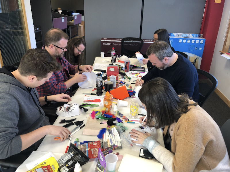 Several workshop participants are designing their own masks. The masks are white and made from paper and they are using different coloured pens, paper and feathers to decorate them.